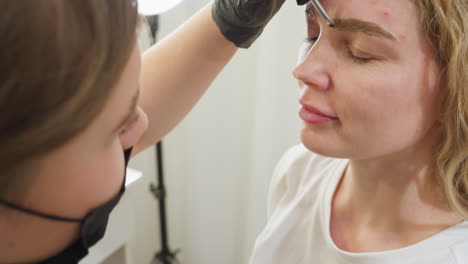 beautician wearing black gloves holds small glass jar containing cream used for eyebrow treatment while preparing to apply on client. blurred background shows part of client and ring light stand