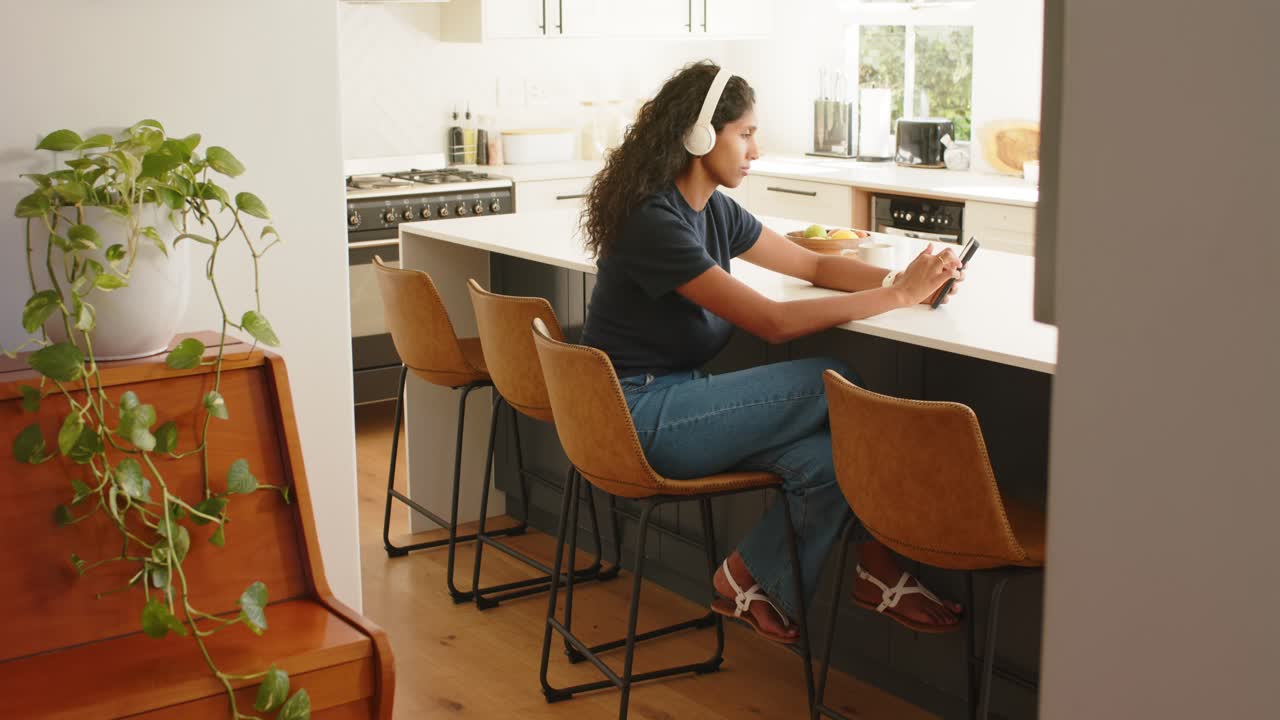 African American woman using smartphone at kitchen island, wearing headphones, reacting to text