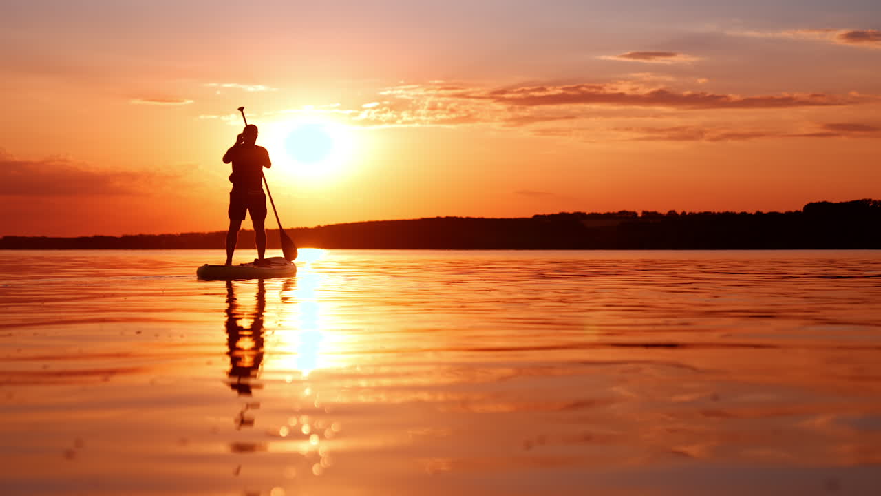Silhouette maschile si muove sull'acqua su tavola da paddle. Lo sportivo rema con un remo al tramonto. Inquadratura dal basso.