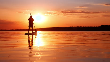 Male silhouette moves by the water on paddle board. Sportsman rows with one oar at sunset. Low angle view.