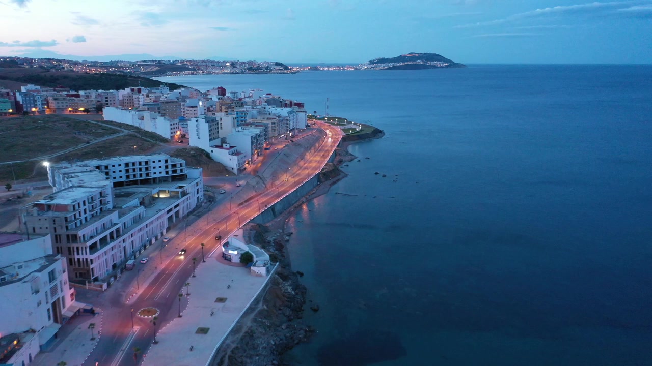 Veduta aerea di una città costiera nel Marocco settentrionale all'ora blu, che mostra una strada lungomare illuminata, moderni edifici sul mare, acqua calma del Mediterraneo e un promontorio distante lungo la costa.