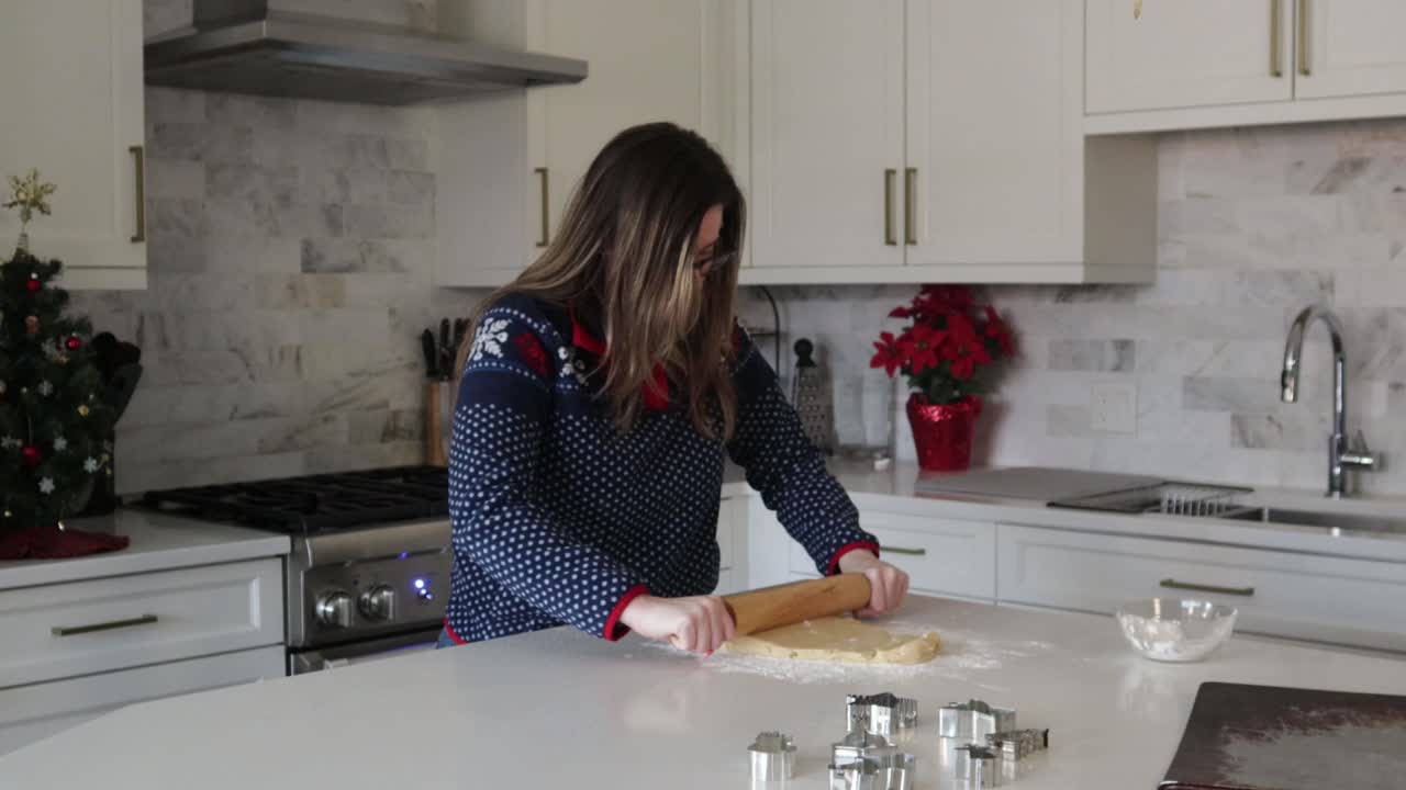 Una donna stende e appiattisce la pasta frolla con un matterello in una cucina luminosa. La cucina è decorata per Natale e ha tagliabiscotti natalizi per preparare biscotti di Natale.