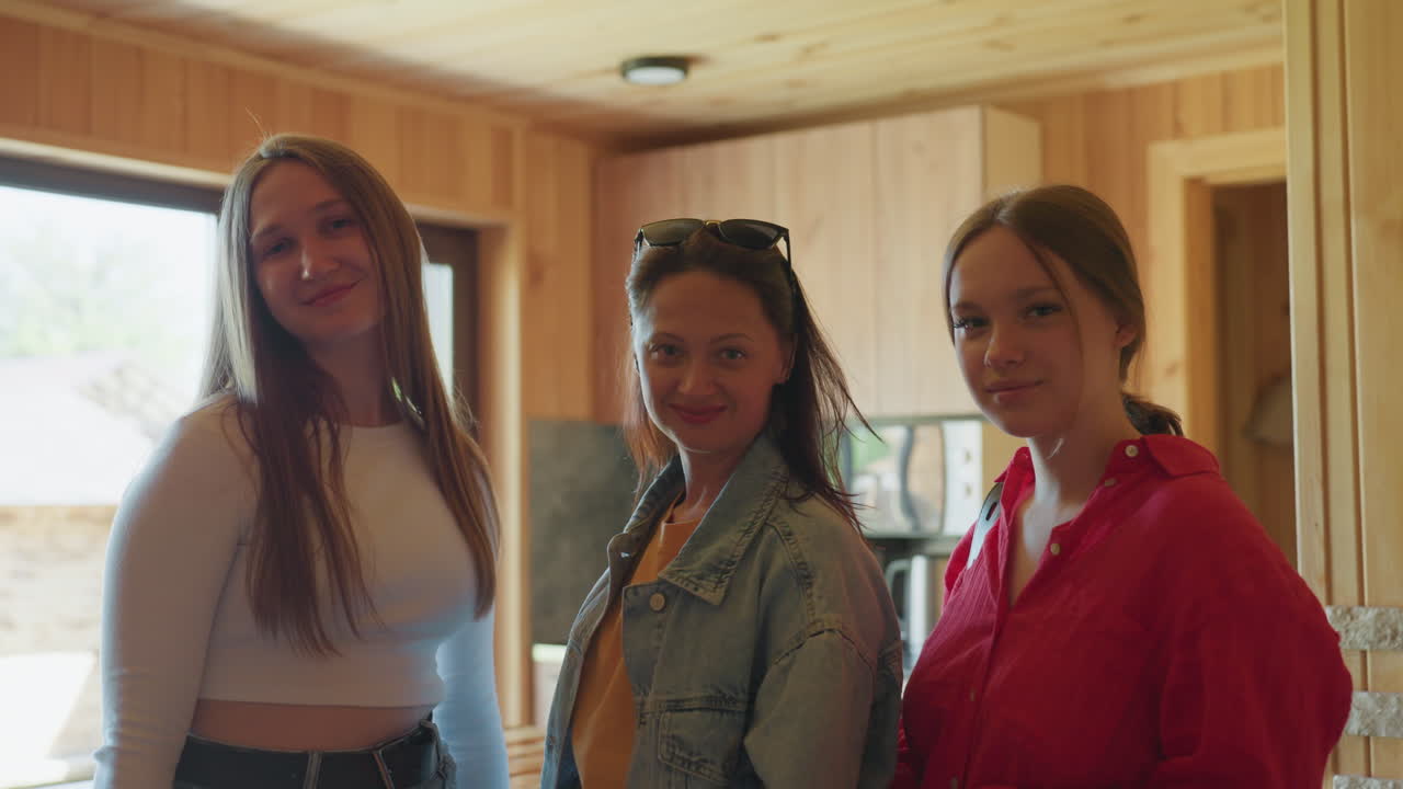Three women stand indoors in wooden interior filled with warm light as woman in middle removes sunglasses with confident gesture while others pose casually beside her near fireplace