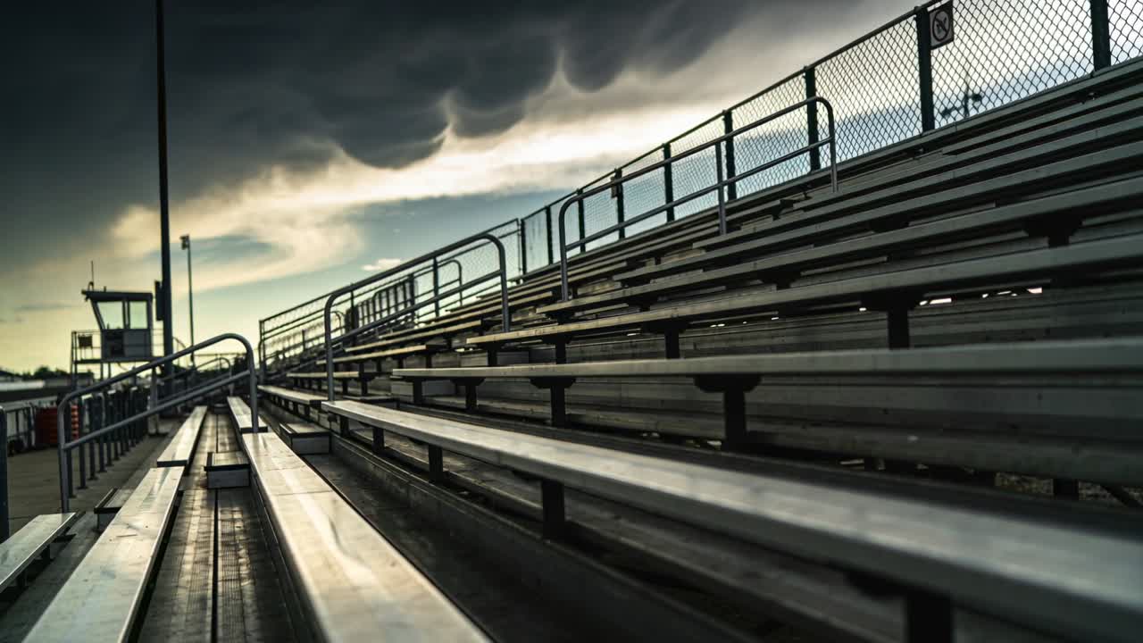 Verfolgende Kamera zeigt zurückweichende Reihen von Metalltribünen in einem kleinen Stadion unter stürmischem Himmel.