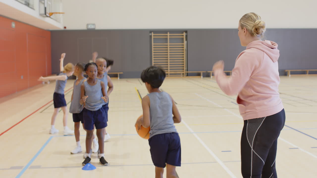 In de schoolgymzaal basketballen kinderen terwijl de coach toekijkt en een bal vasthoudt.