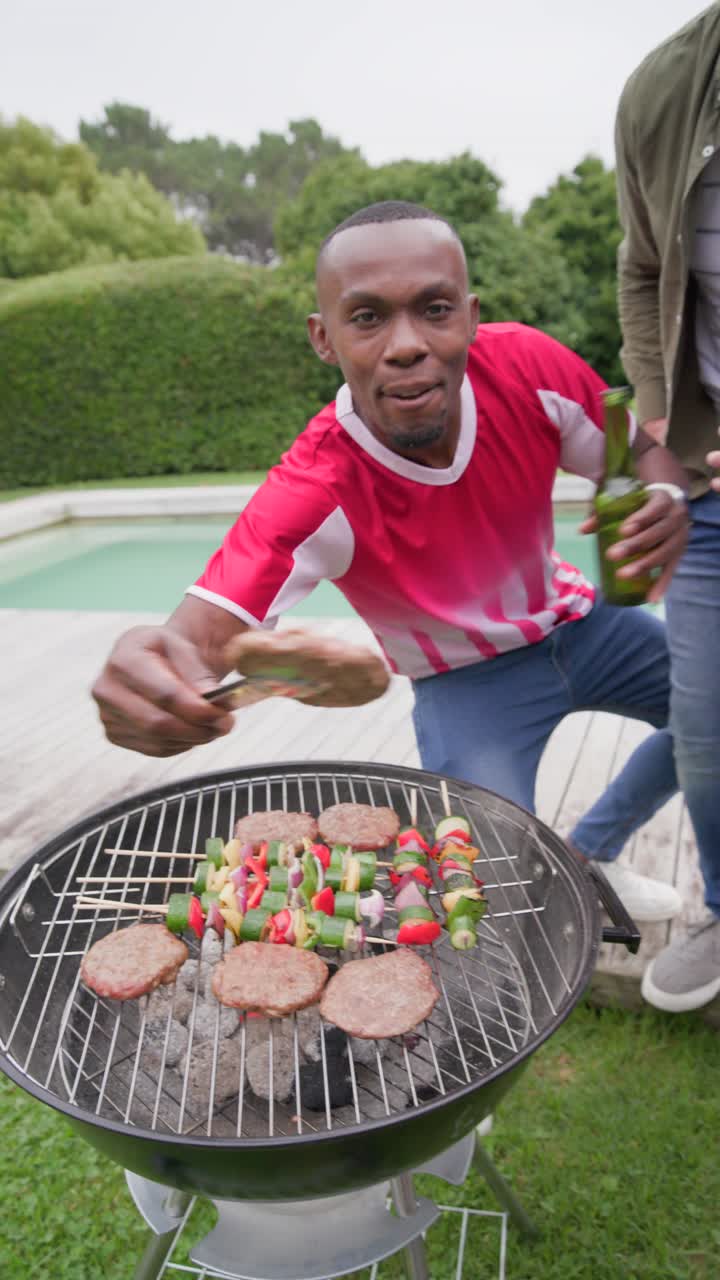 Video verticale: Uomini di diverse etnie che si scattano un selfie con birre verdi su un barbecue in terrazza, mentre uno griglia con una spatola.