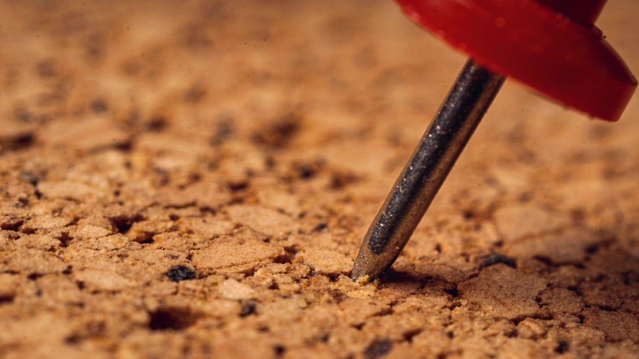Pushing metal tip of red pushpin into corkboard on desk, releasing cork fragments, copy space