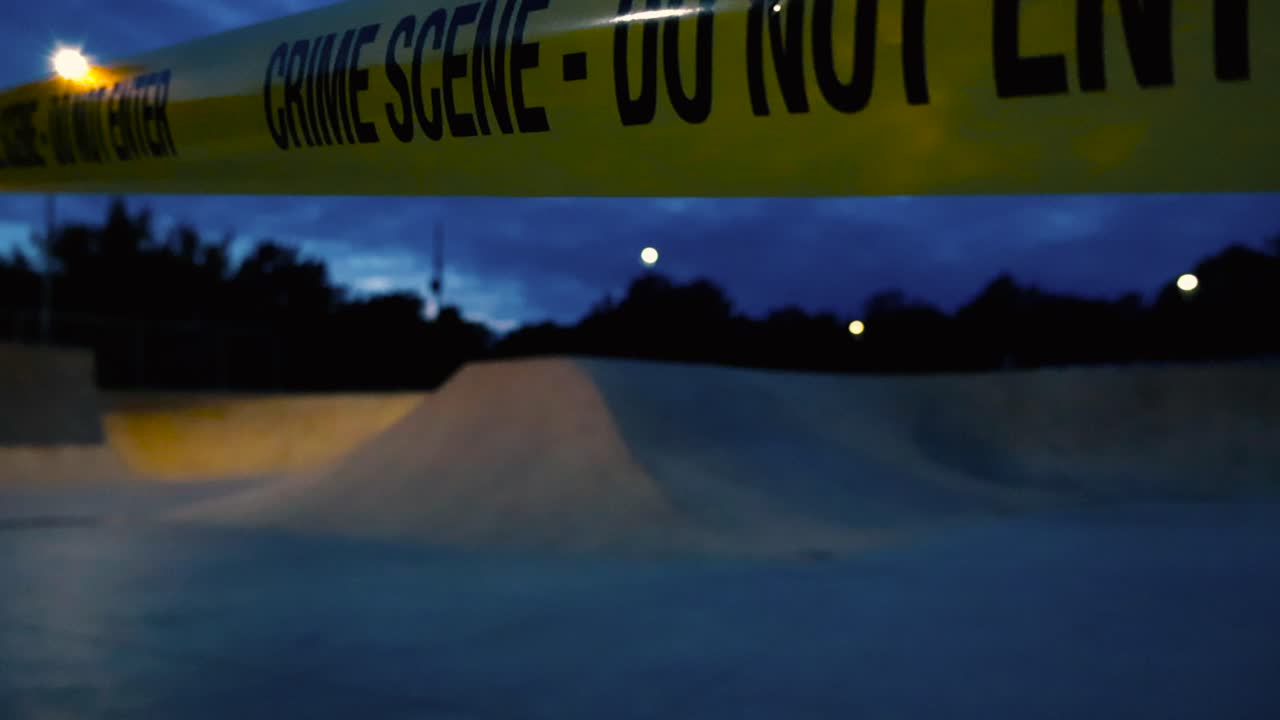 Low angle close up or close up video of a yellow vibrant colored Crime Scene Do Not Enter police tape ribbon pulled in front and around a evening skatepark while street lights are in the background
