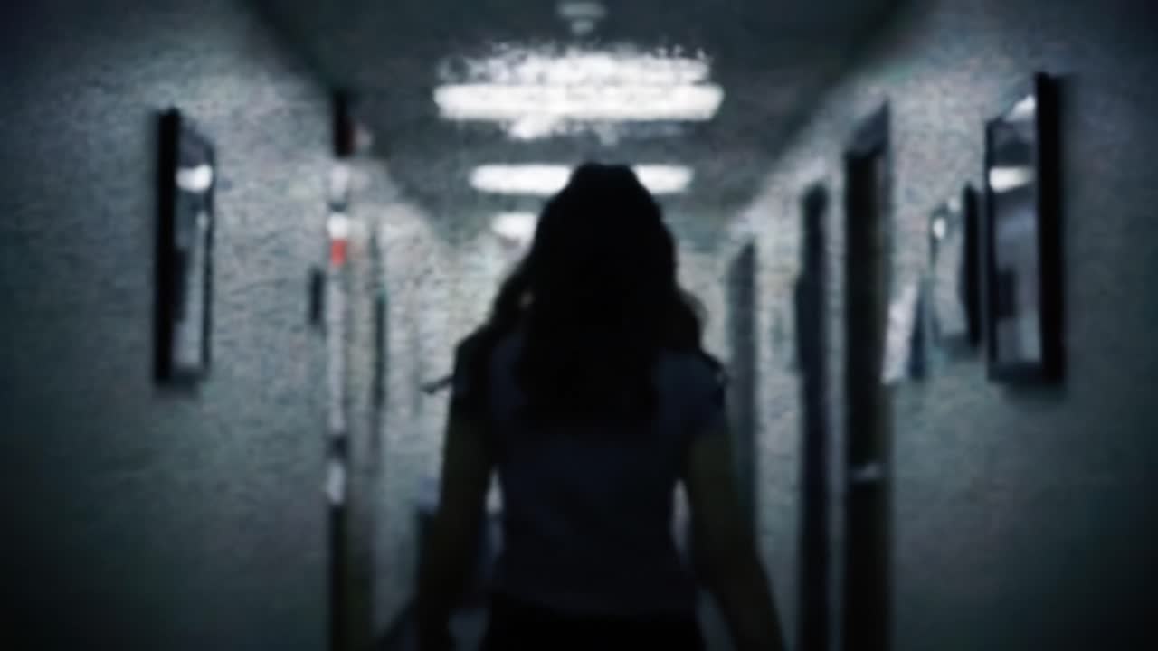 A person walks down a dimly lit hospital corridor, creating a tense atmosphere filled with anticipation and mystery. Shadows loom as they navigate the unsettling environment.