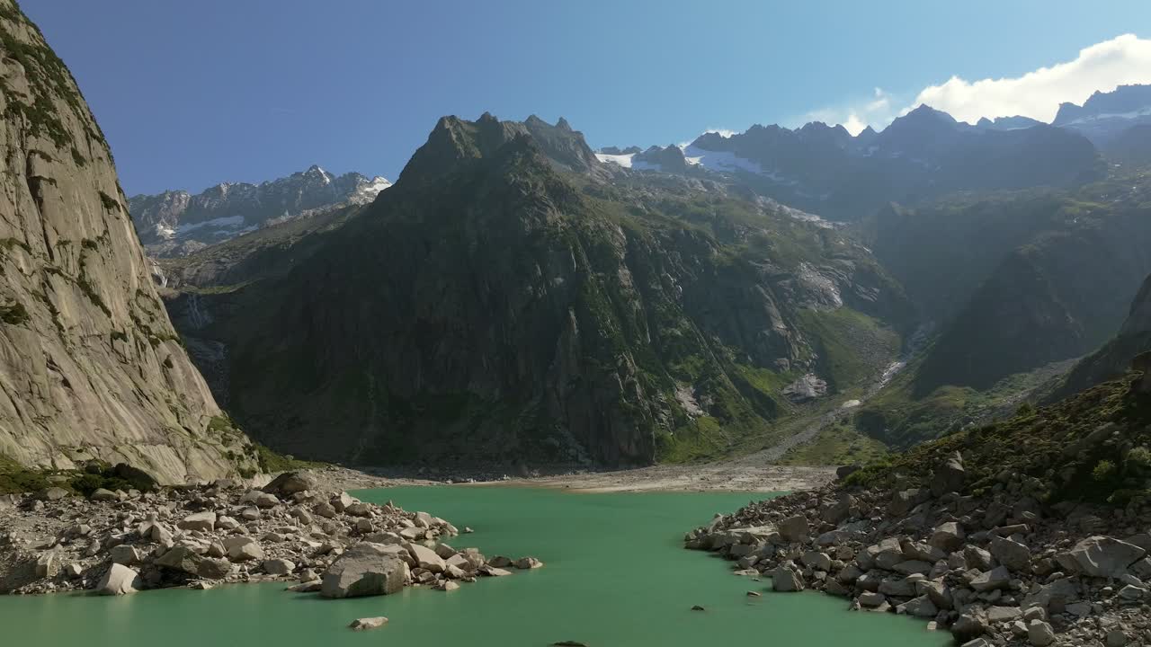 Aerial shot over the Gelmersee Lake, showing turquoise alpine waters surrounded by steep rocky cliffs and dramatic mountain peaks under a clear summer sky
