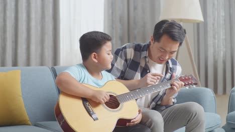 asian father teaching his son about guitar chords on sofa at home