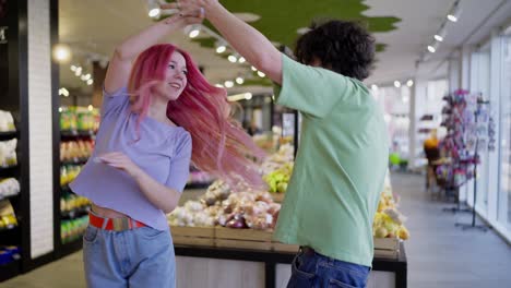 Happy-girl-with-pink-hair-and-brunette-guy-in-a-green-T-shirt-dancing-in-a-grocery-store.-Happy-bright-couple-dance-in-grocery-store-while-shopping