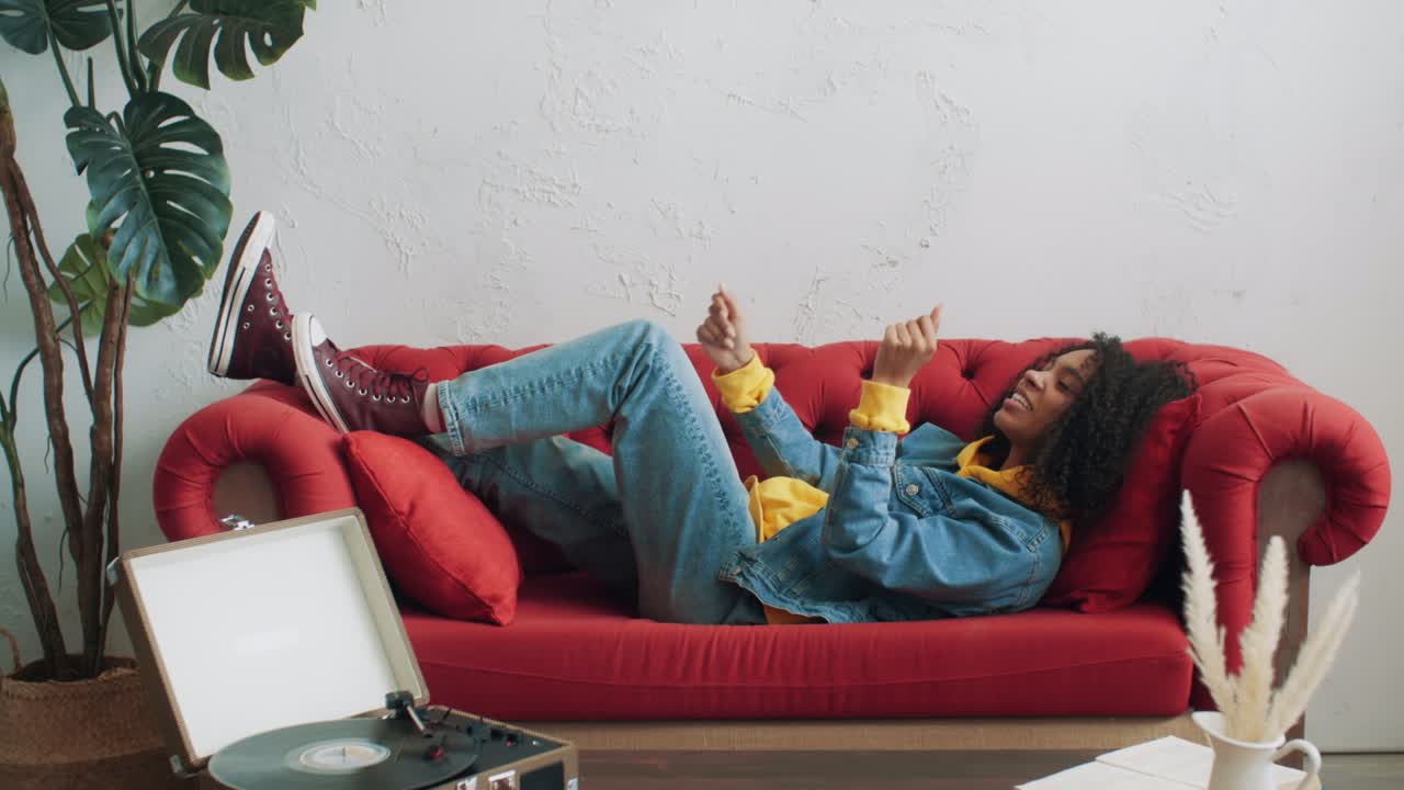 Stylish black female having fun listening music on vinyl record player lying on red sofa. Positive african american urban woman enjoying pastime at home