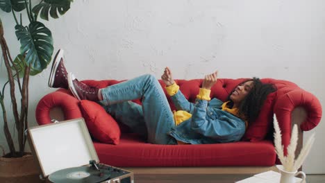 stylish black female having fun listening music on vinyl record player lying on red sofa. positive african american urban woman enjoying pastime at home