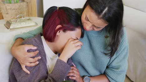 Biracial-sisters-sitting-on-sofa,-crying-and-embracing,-in-slow-motion