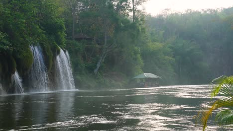 A-beautiful-view-of-floating-houses-on-a-river-and-a-stunning-waterfall-in-the-middle-of-the-jungle-of-Sai-Yok-National-Park-in-Thailand-with-the-pretty-morning-mist-and-exotic-vibes