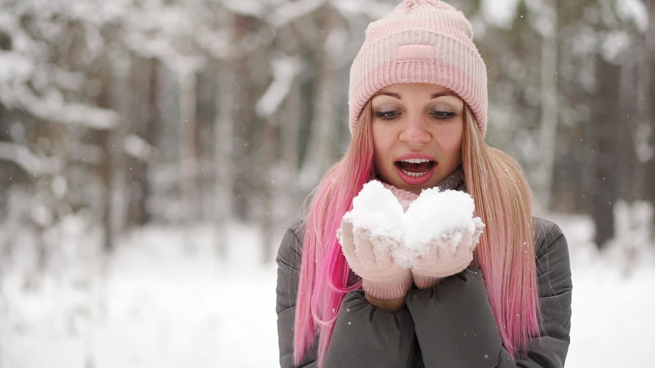 슬로우 모션, 겨울에 재킷 모자와 스카프를 두른 한 여성이 숲 속에서 눈을 손에 쥐고 카메라를 향해 불고 있습니다.