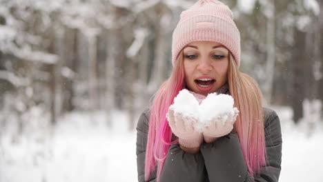 Slow-motion,-a-woman-in-a-jacket-hat-and-scarf-in-the-winter-in-the-forest-holding-snow-in-her-hands-and-blowing-into-the-camera-throws-snow.