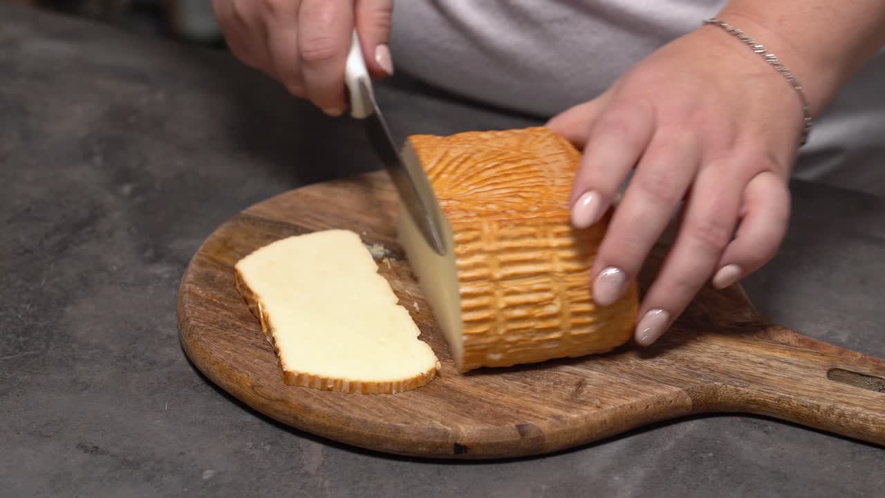 Mão fatiando um bloco de queijo Trapista com uma faca em uma tábua de madeira rústica.