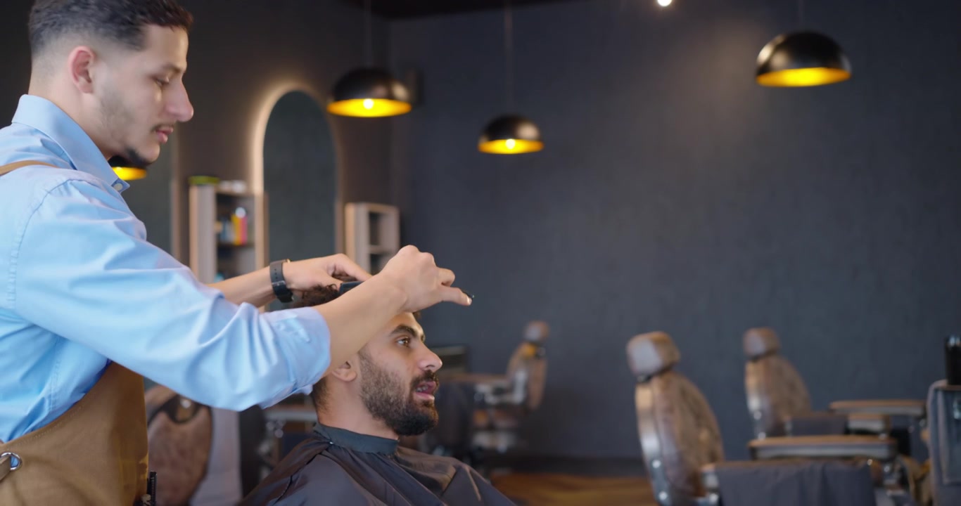 Mandlig barber og siddende kunde, barber retter sort kappe, iført forklæde, forbereder klipning i salonen