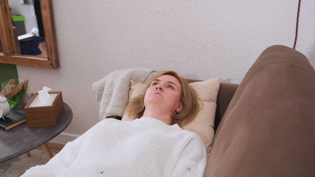 Healing companion lies on brown sofa facing ceiling engaging in conversation with therapist while clock, tissue box, notepad and pencils rest on round table beside cozy session setup