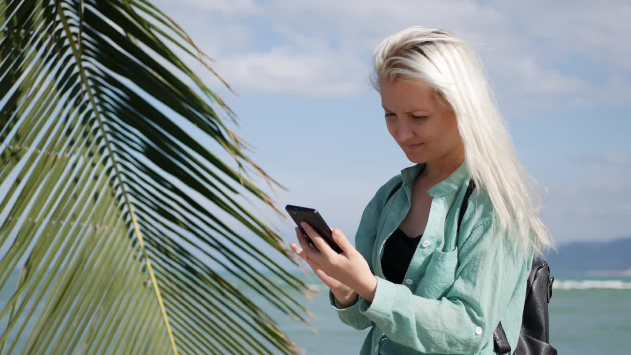 hermosa mujer delgada con cabello rubio largo y camisa verde de pie cerca de la palmera y usando el teléfono inteligente sobre el fondo del mar. niña pantalla táctil
