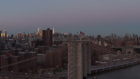 American-flag-waving-in-wind-on-top-of-bridge-tower.-Cityscape-with-high-rise-glossy-buildings-reflecting-colourful-twilight-sky.-Manhattan,-New-York-City,-USA