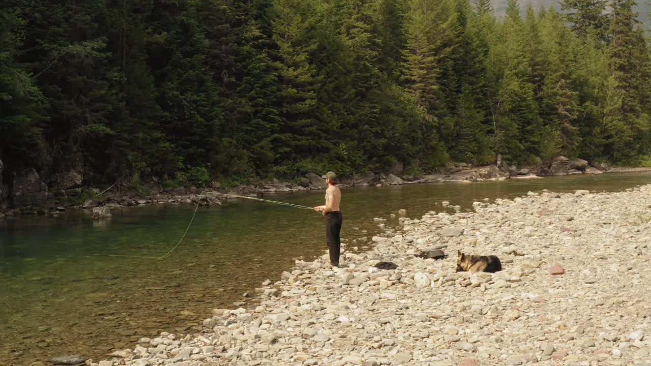 pesca con mosca en mcdonald creek en el parque nacional de los glaciares - un hombre echando moscas mientras su perro leal espera pacientemente en el banco