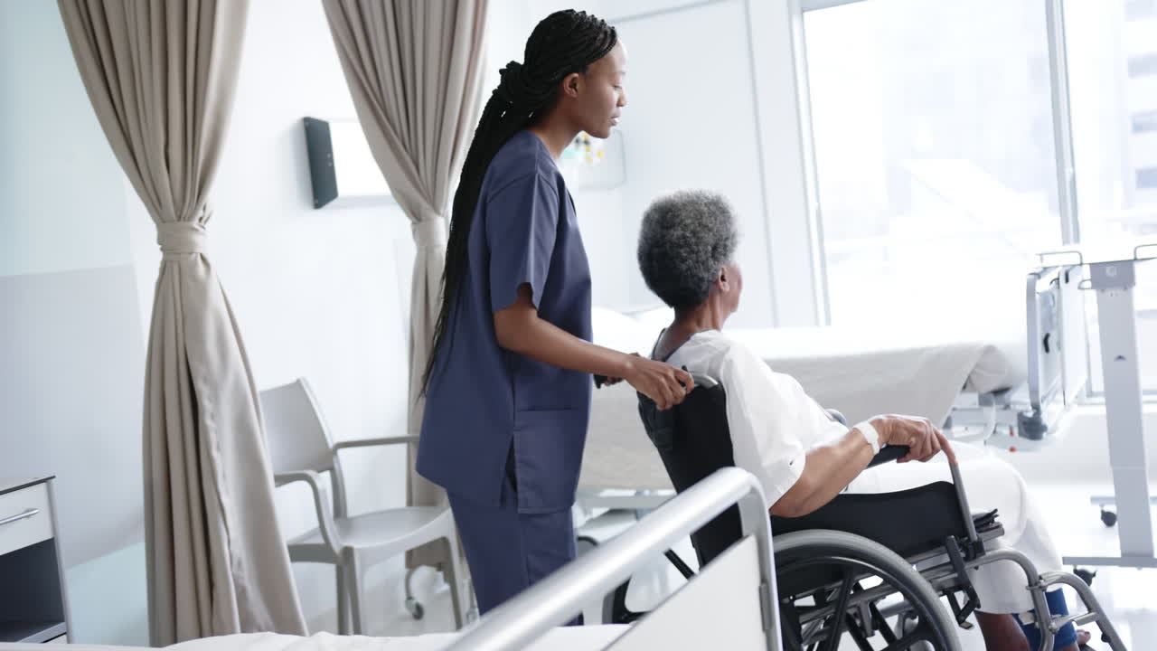 African american female doctor talking with senior woman in wheelchair in hospital room, slow motion