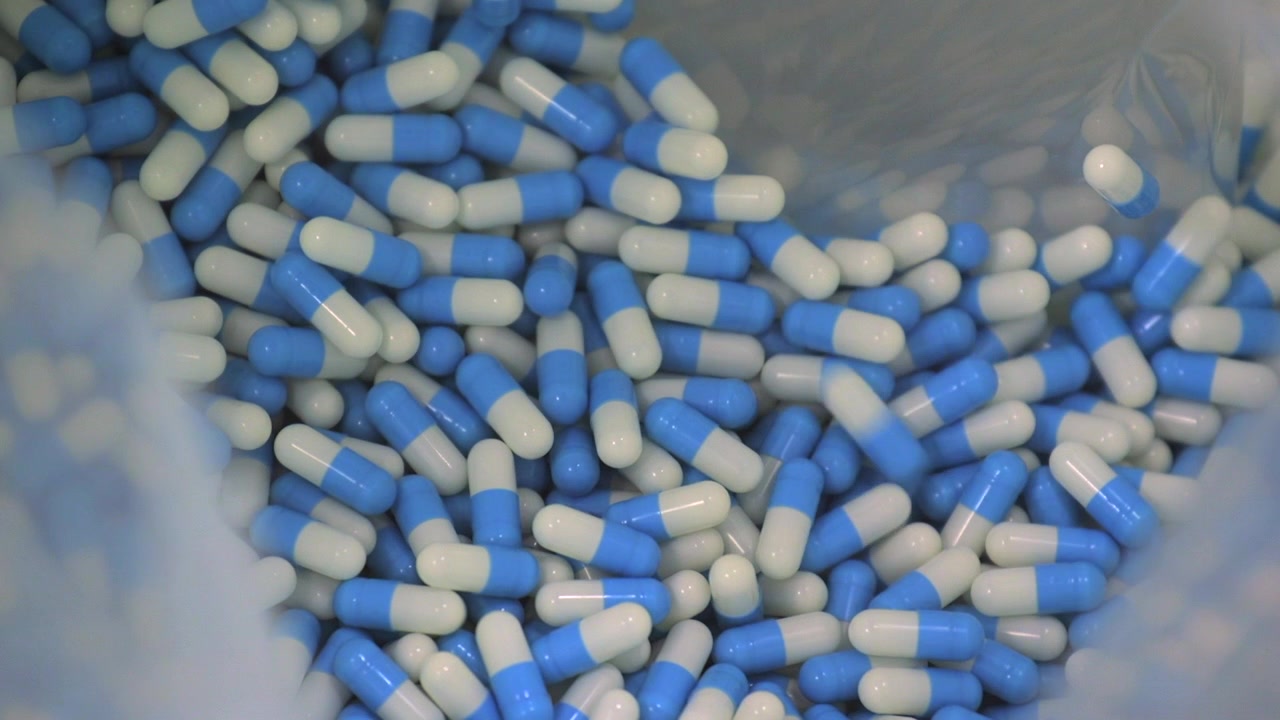 Capsule farmaceutiche blu e bianche cadono da una macchina di produzione automatizzata in un contenitore di raccolta all'interno di un moderno impianto di produzione farmaceutica.