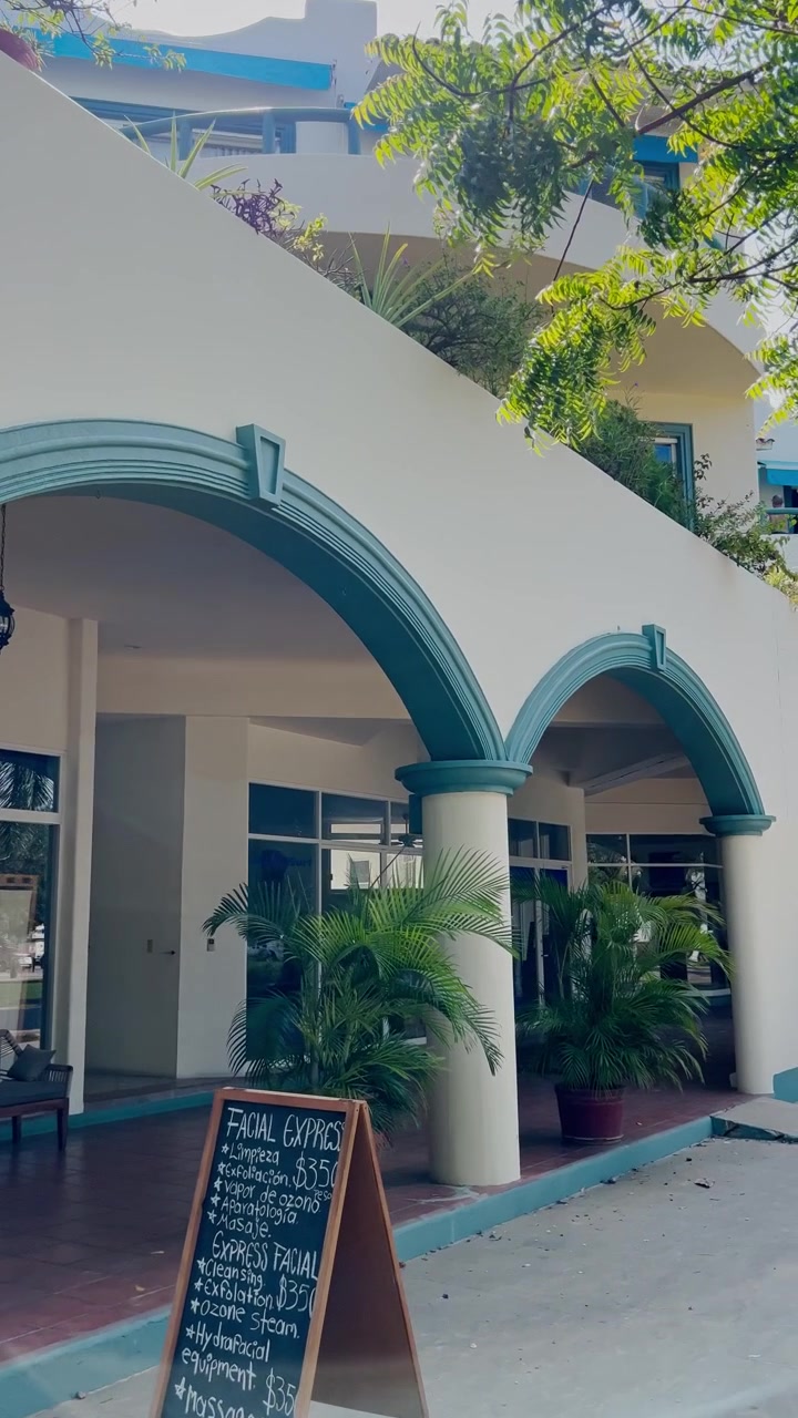 Edificio de estuco blanco con entradas arqueadas azules, columnas redondeadas, puertas de cristal y plantas tropicales a lo largo del pasillo exterior a plena luz del día.