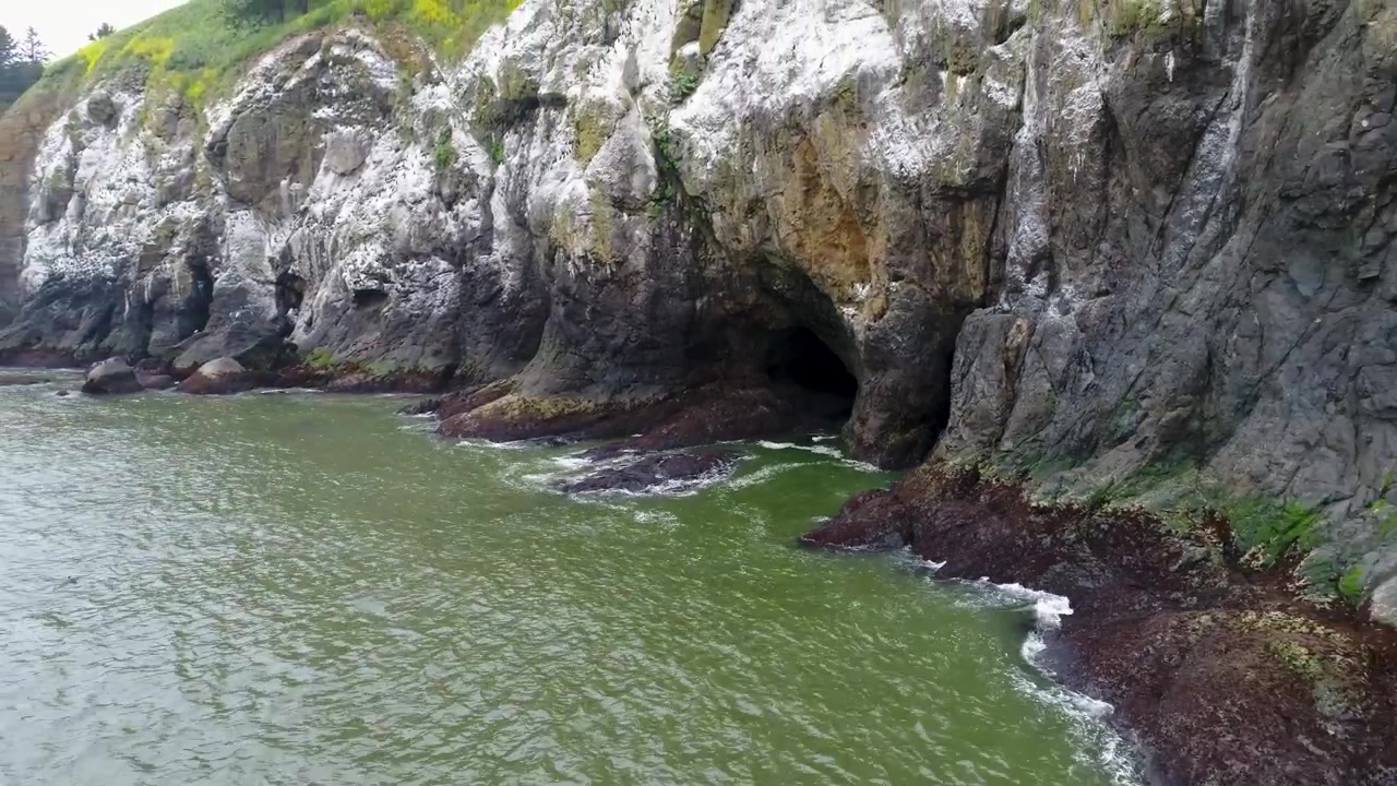 A cinematic reversing drone shot, capturing a sea cave entrance in the side of a cliff, near Deadman's Cove at Cape Disappointment, where the Columbia River meets the Pacific Ocean in Washington.