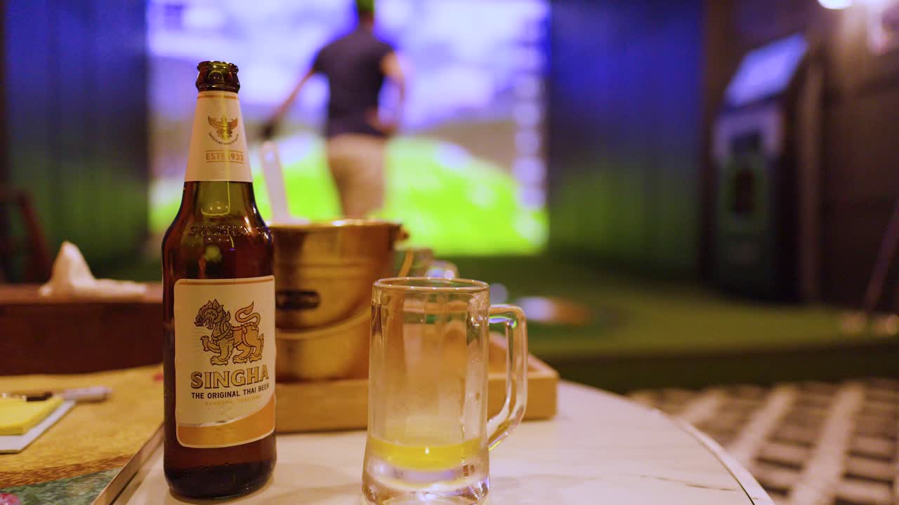 Una bottiglia di birra e un bicchiere sono su un tavolo mentre un golfista esegue uno swing sullo sfondo.