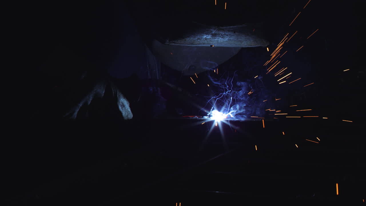 A blacksmith with a welding mask and welders leathers, is welding metal products in the garage, fire sparks fly to the sides. Dark mood. Slow Motion shot with Red Monstro 8K.