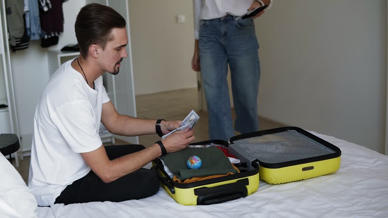 бородатый человек сидит на кровати, готовясь к отдыху - нам долларовые банкноты - человек подсчитывает чемодан с одеждой, когда его