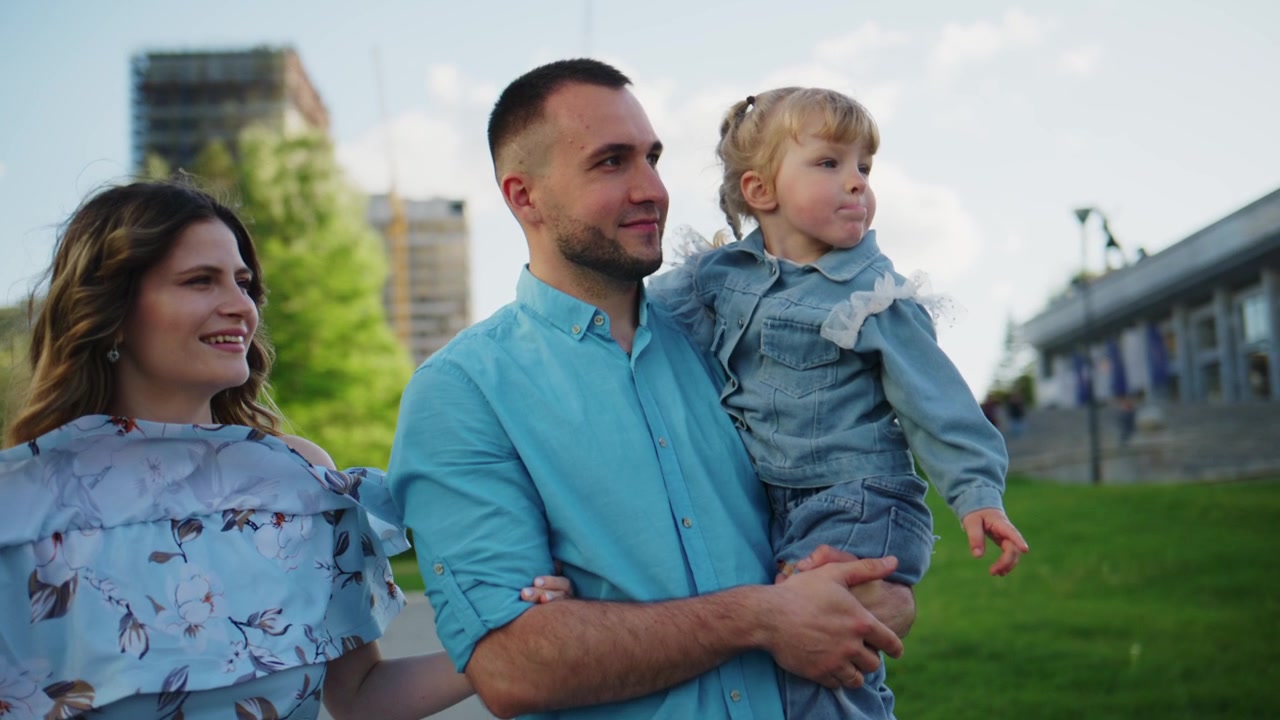Familia feliz de tres disfrutando de un momento al aire libre juntos. La madre sonríe cálidamente junto al padre que sostiene a su hija pequeña. La niña mira con curiosidad, explorando los alrededores