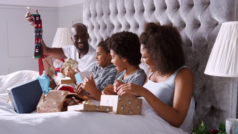 young mixed race family sitting up in bed together unwrapping presents on christmas morning, side view, close up