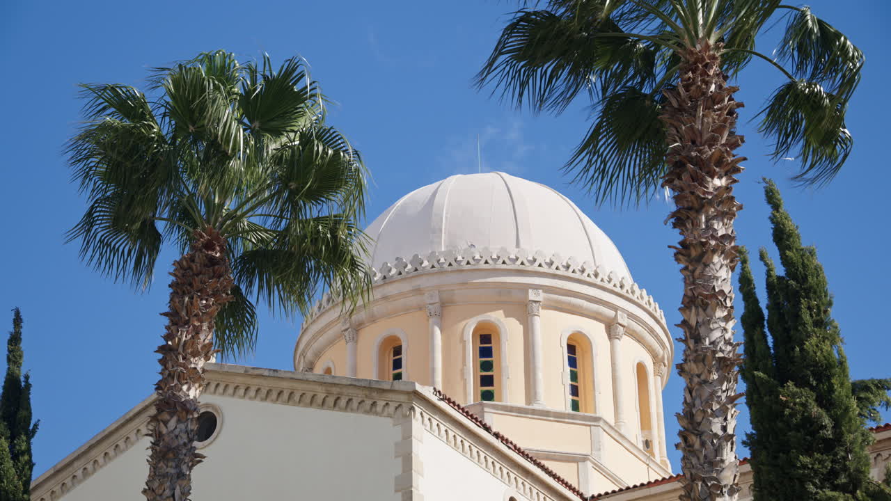 Pemandangan detail kubah gereja yang dibingkai oleh pohon palem tinggi di Limassol, Siprus. Arsitektur keagamaan Mediterania tradisional di bawah sinar matahari cerah dan langit biru