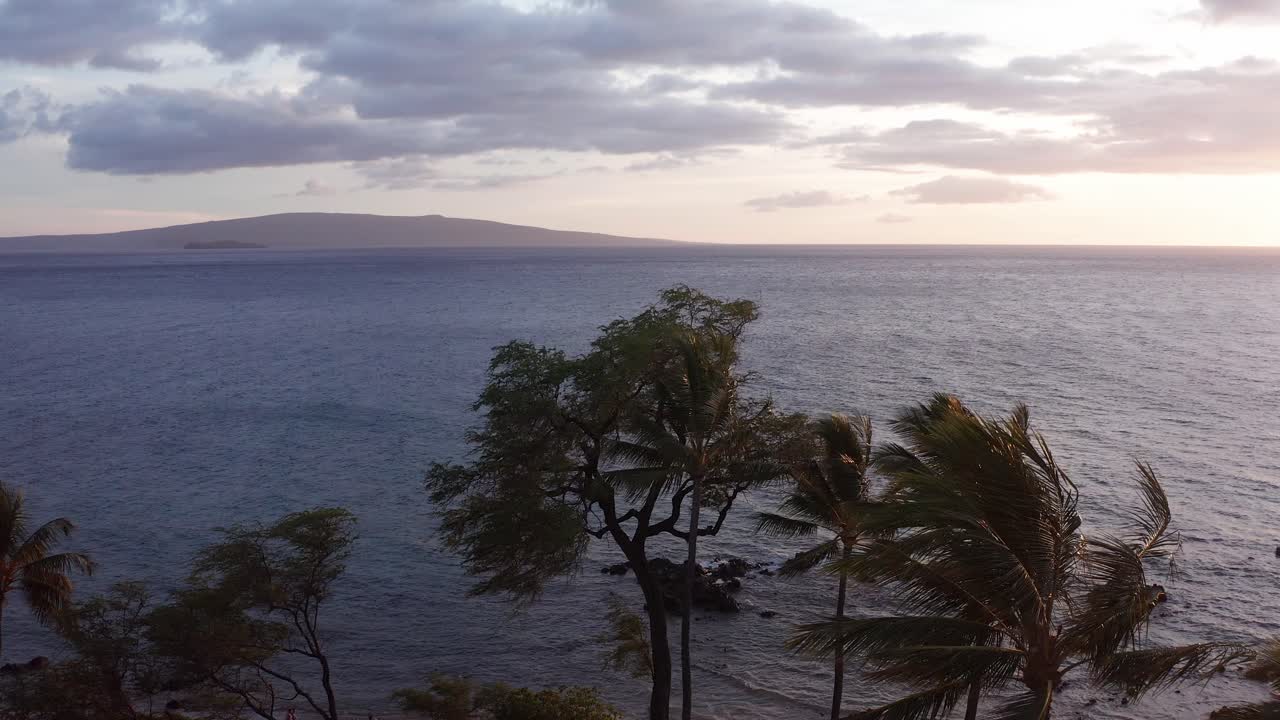 Low and slow aerial shot flying over pam trees along the beach in Wailea with Molokini Crater and the sacred island of Kaho'olawe in the distance at sunset in Maui, Hawai'i