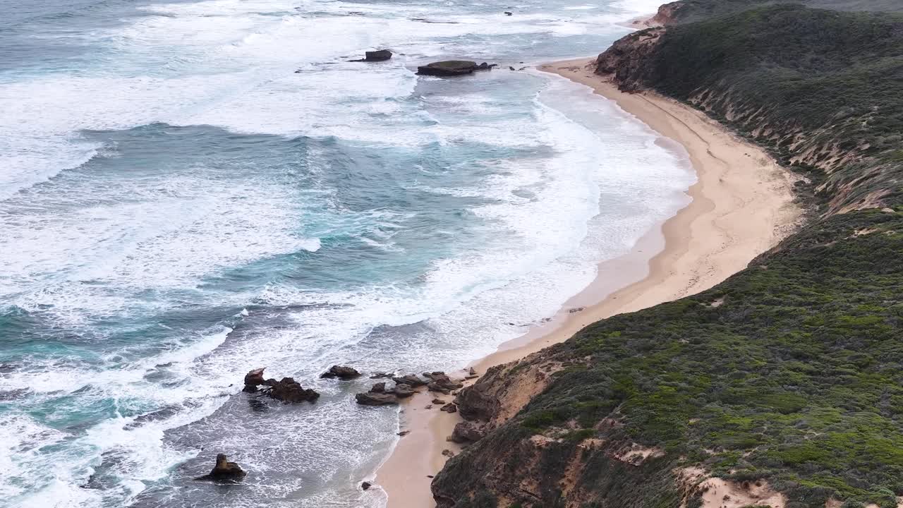 Drone pans above Portsea coastline, revealing cliffs, sandy beach, ocean waves, and lush vegetation
