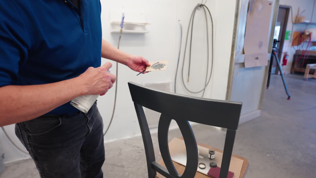 Slow-motion close-up as blue-shirted worker applies clear coat to grey chair