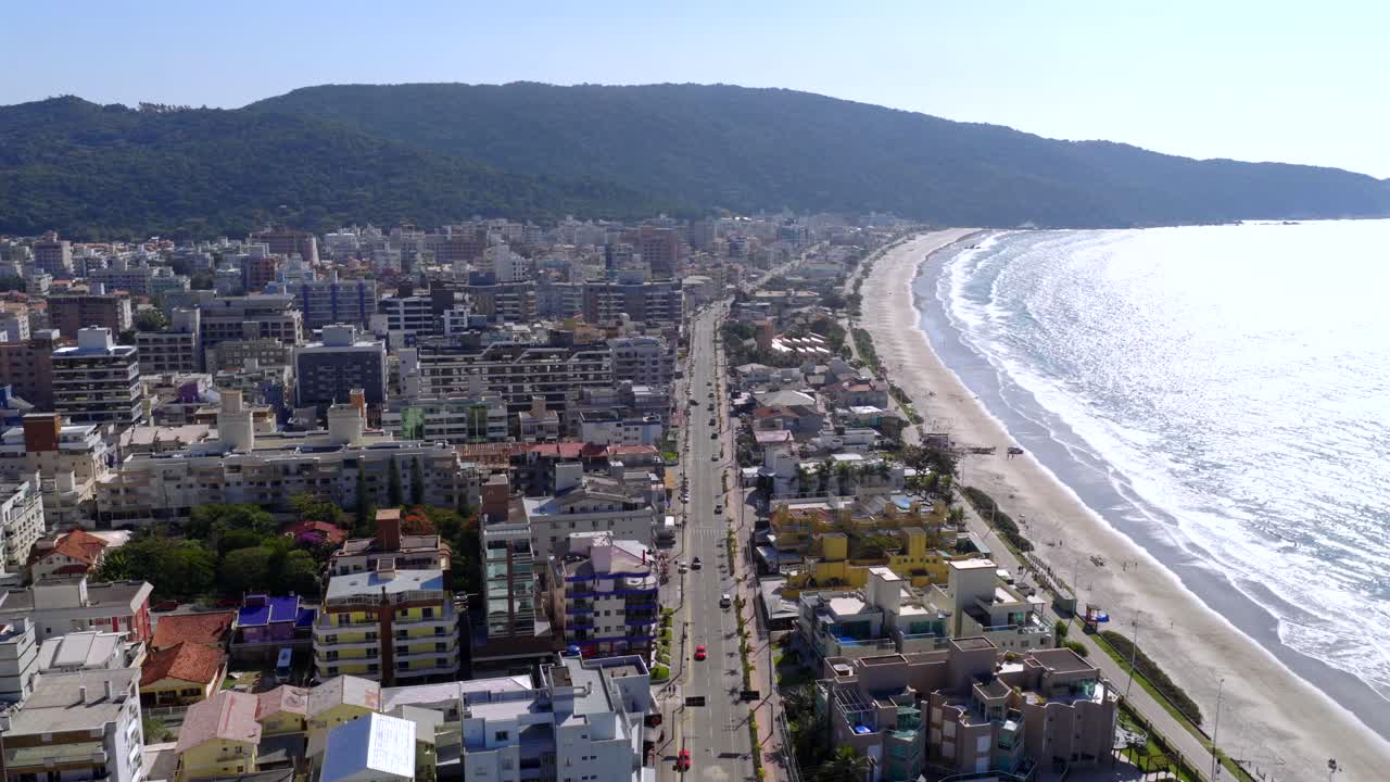 Drone glides parallel to Bombinhas beach during ocean early afternoon. Bright light