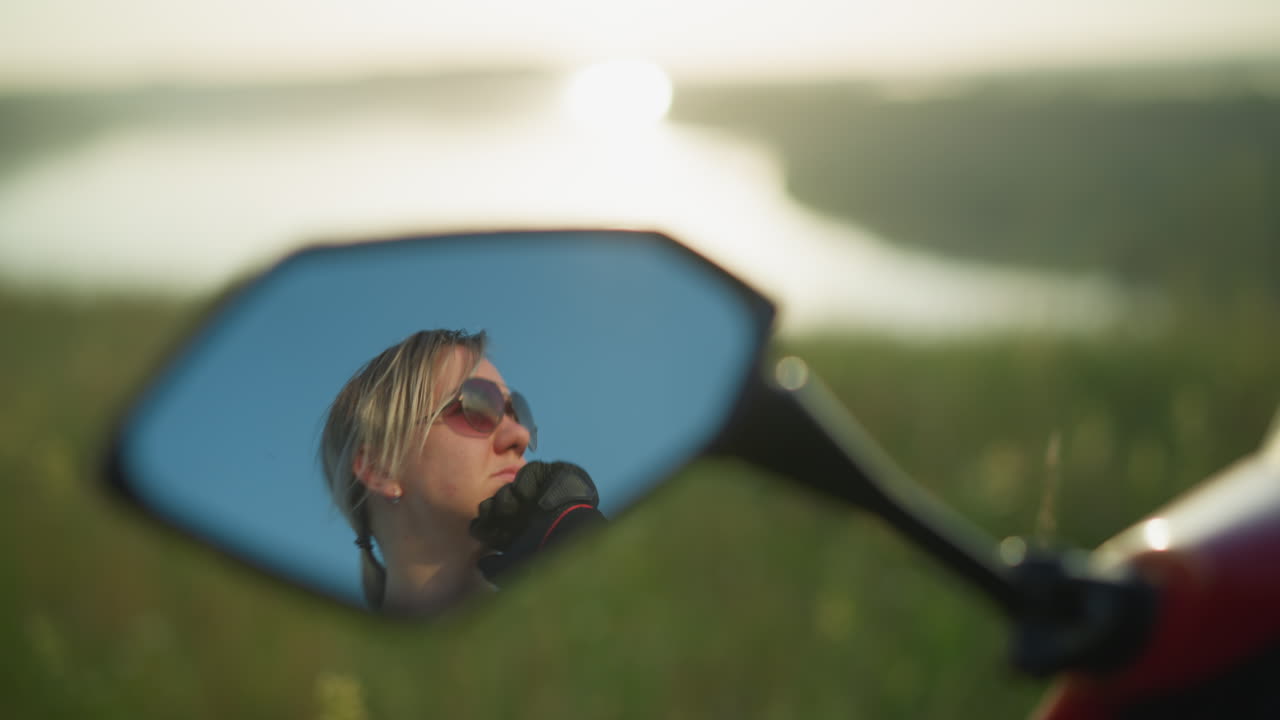 Mirror reflection of a woman wearing sunglasses, resting her chin on her hands while gazing intently at something, the background shows a soft blur of a field and water