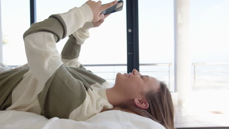 lying on bed, young woman using smartphone and smiling in home setting, copy space