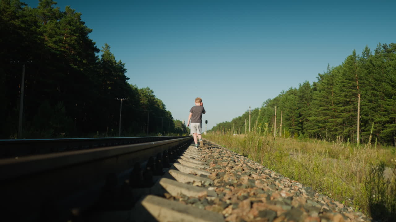 Junge, der allein auf einer Bahntrasse geht, umgeben von Wald und Strommasten, wobei frühes Sonnenlicht einen warmen Schein über die ruhige Landschaft wirft