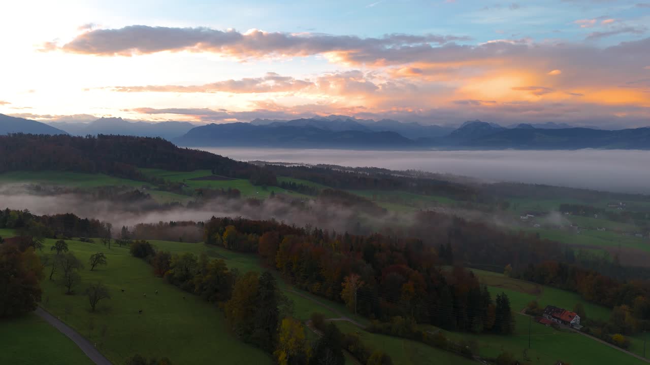 Drone flight over an autumn landscape in Switzerland. The sea of   fog and the hilly landscape is illuminated at sunrise. In the background you can see the mountain ranges. The clouds glowing orange.
