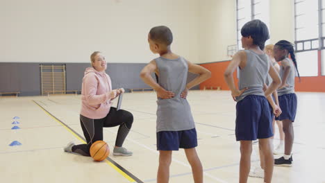in school gym, coach kneeling with basketball, instructing students during practice