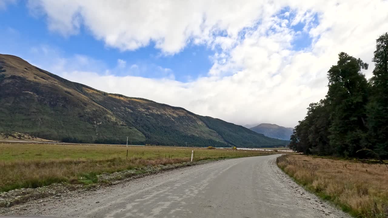 Vehicle travels down rural gravel road, passing fields, trees, and mountains under partly cloudy sky