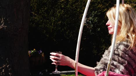 Attractive-woman-enjoys-glass-of-wine-in-garden-chair-swing-wide-4k-slow-motion