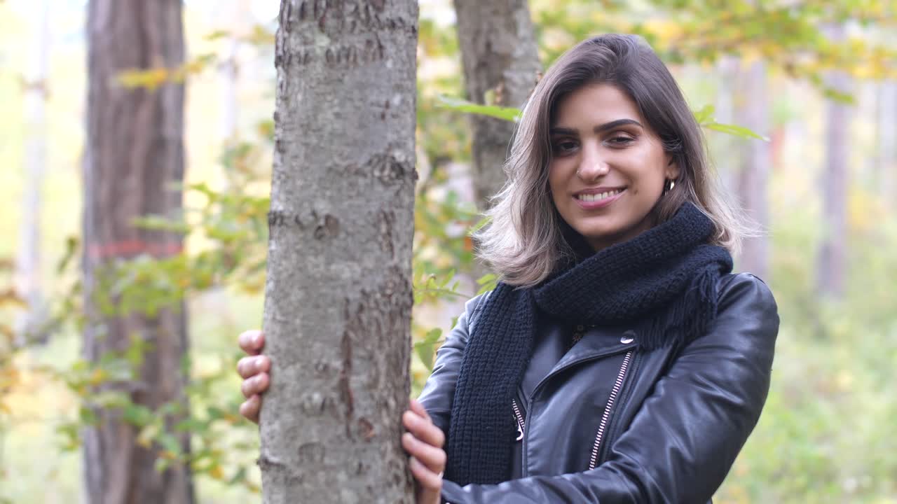 chica morena en el bosque de otoño se encuentra junto a un árbol y sonríe a la cámara